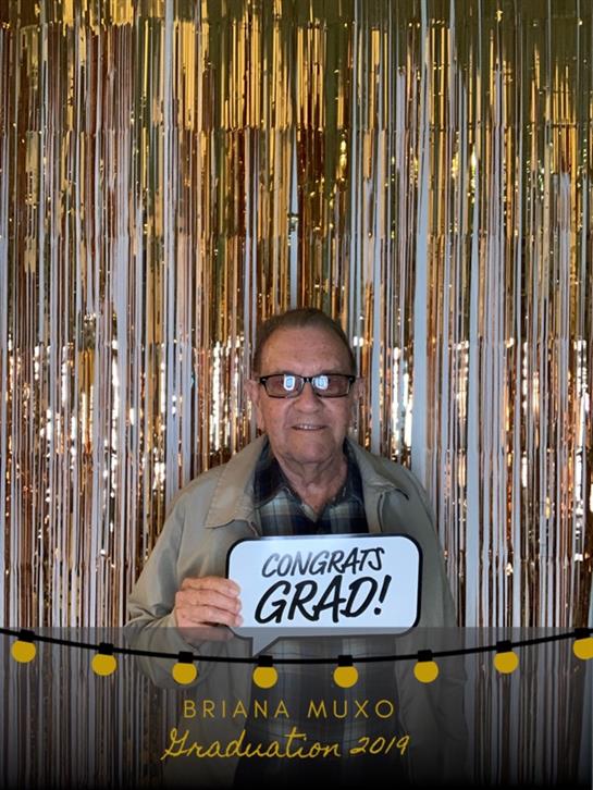 An elder man joyfully holds a congrats sign at a graduation celebration surrounded by shiny decor.