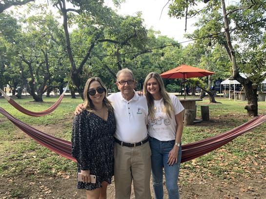 Two women and an older man smile together in a park, surrounded by trees and hammocks.