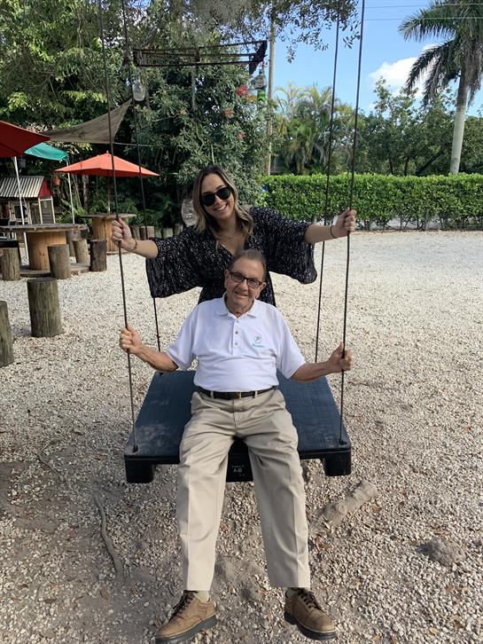 A young woman pushes an elderly man on a swing in a sunny park filled with greenery.