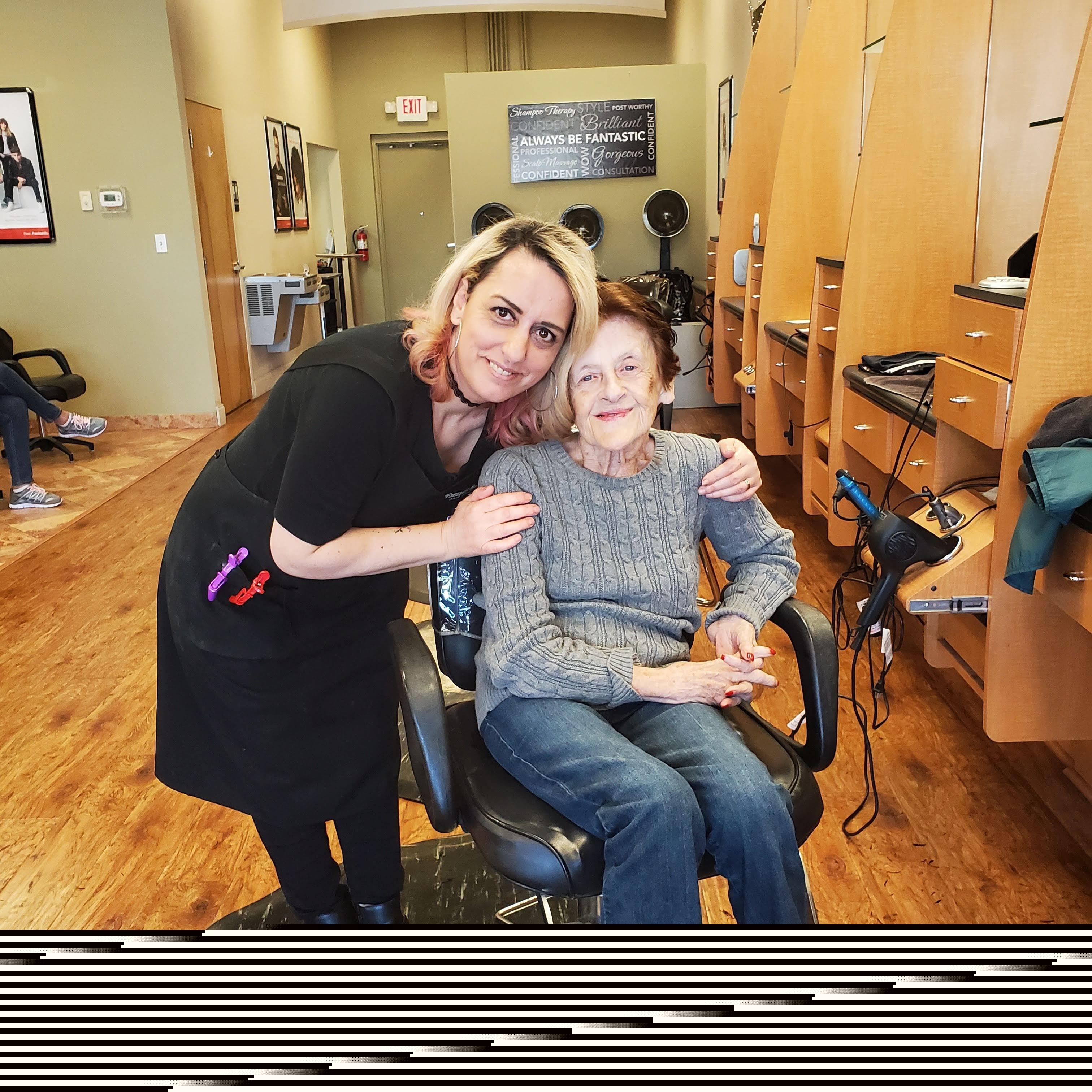 A hairstylist smiles alongside an elderly client seated in a salon chair during a makeover session.