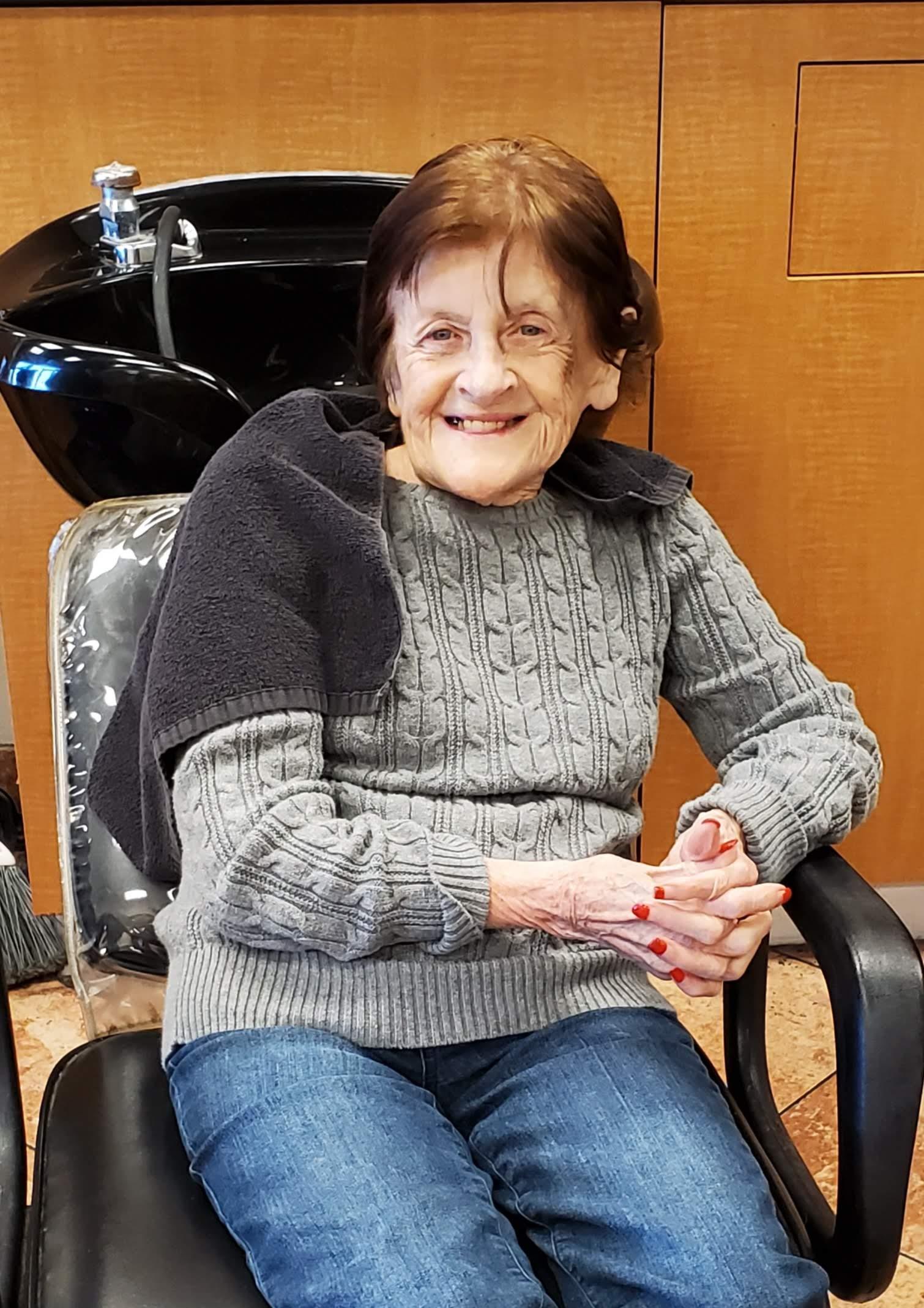 An elderly woman relaxes and smiles in a salon chair before her haircut appointment.