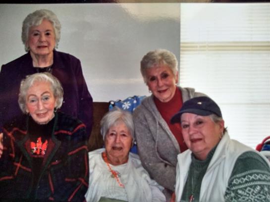 A group of five older women share laughter and stories in a warm living room setting.