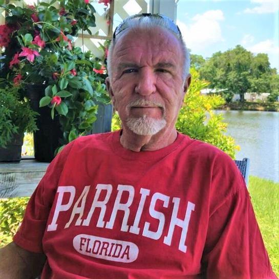 Elderly man relaxes outside in Florida, wearing a red shirt, with lush greenery and water in view.