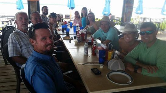 Friends gather around a table filled with food and drinks at a beachside location during daytime.