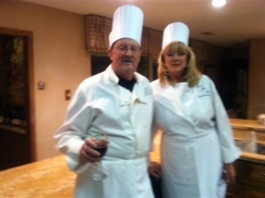 Two chefs in white uniforms stand together, one holding a glass of wine, in a warm kitchen.
