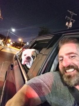 A man smiles joyfully while driving with his dog in the car at night on a quiet street.