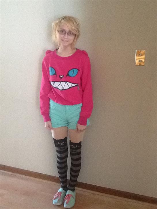 A young person wearing a bright pink sweater and blue shorts poses confidently indoors.