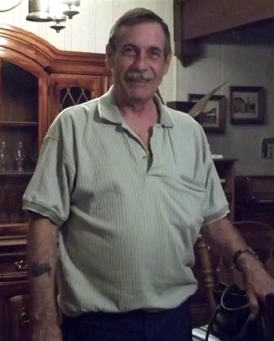Man stands smiling in a warm living room with wooden furniture and personal items visible.