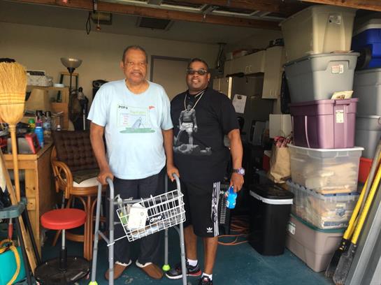 Two men chat in a cluttered garage, one with a walker and the other holding a drink.