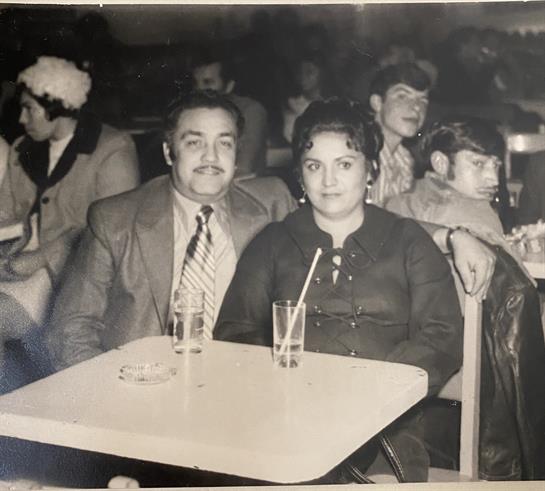 A couple sits together at a table, enjoying drinks while others socialize in the background.
