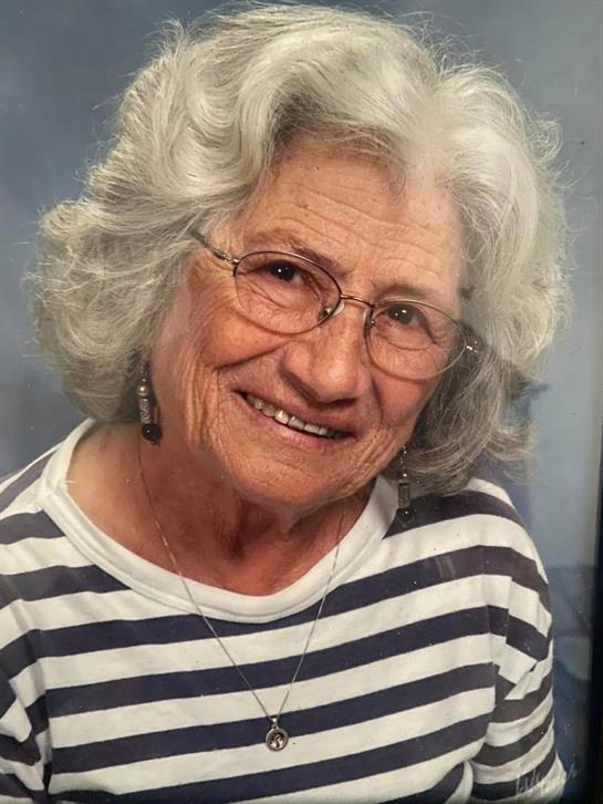 An elderly woman with gray hair smiles warmly while wearing a striped shirt and glasses.