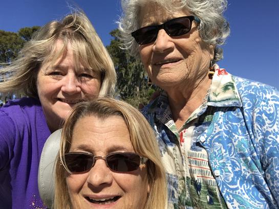 Three women smile at the camera, wearing sunglasses and enjoying a sunny day at the park.