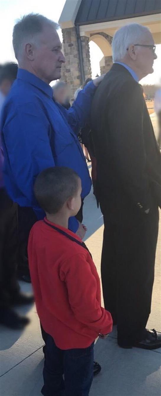 Individuals gathered around a child in bright clothing during an outdoor gathering in sunlight.