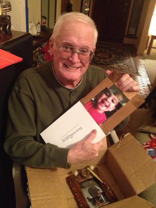 An elderly man smiles while holding a card that features a child's photo during a family gathering.