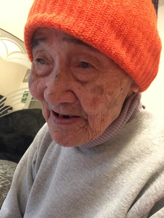 An elderly woman with a joyful expression wears an orange hat while sitting inside her home.
