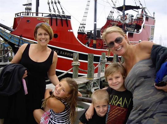 Two women and three children smile as they pose by the dock next to a vibrant red pirate ship.
