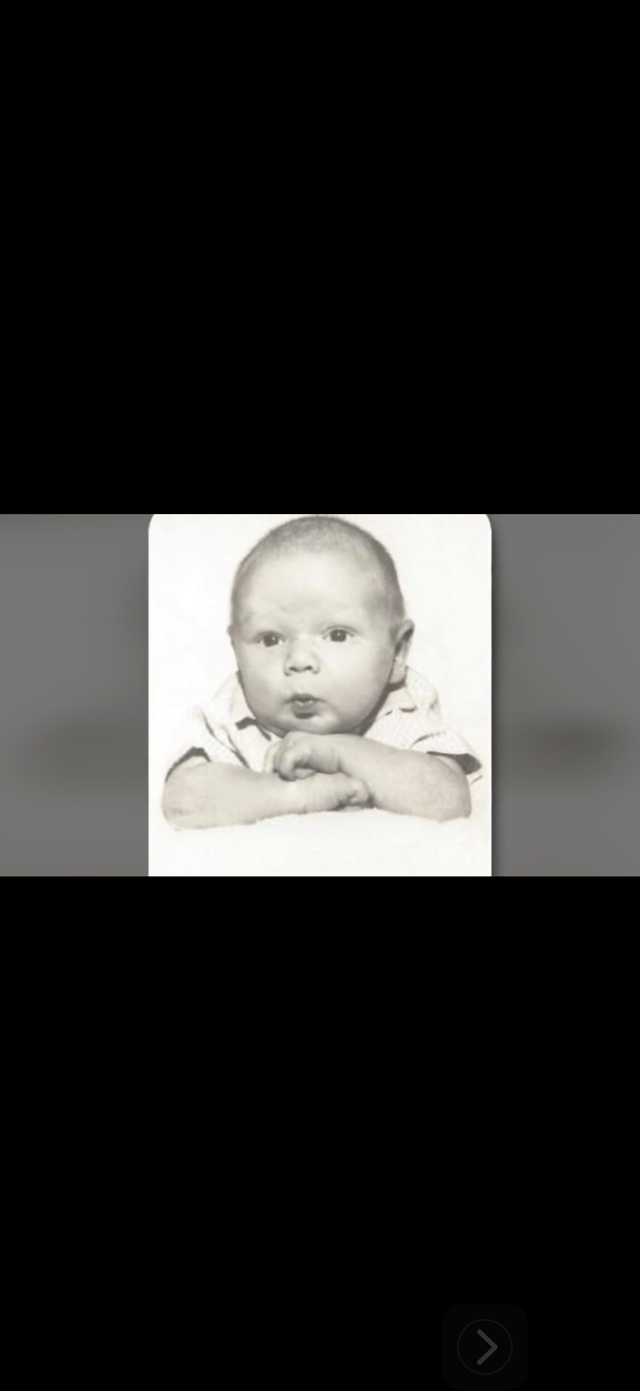 A baby with a curious expression lies on their stomach, hands clasped under their chin.