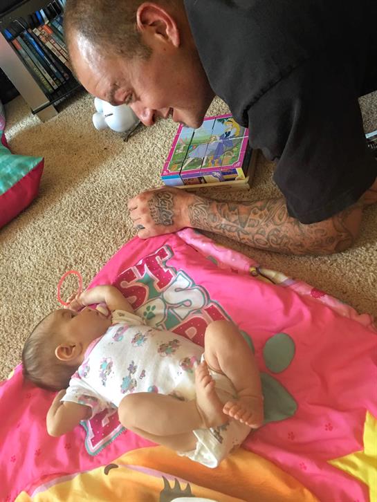 A father plays on the floor with his baby on a colorful blanket full of toys.