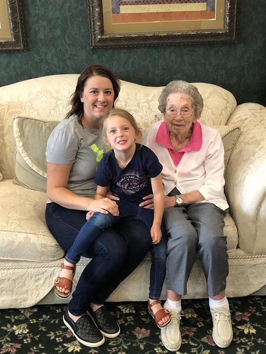 A young girl sits on a couch with her mother and grandmother, all smiling happily together.