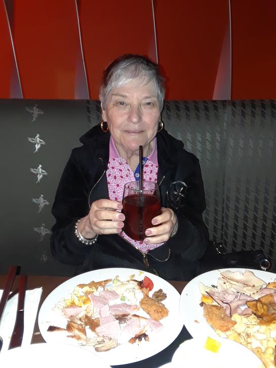 An elderly woman smiles at a restaurant table, drink in hand and food around her.