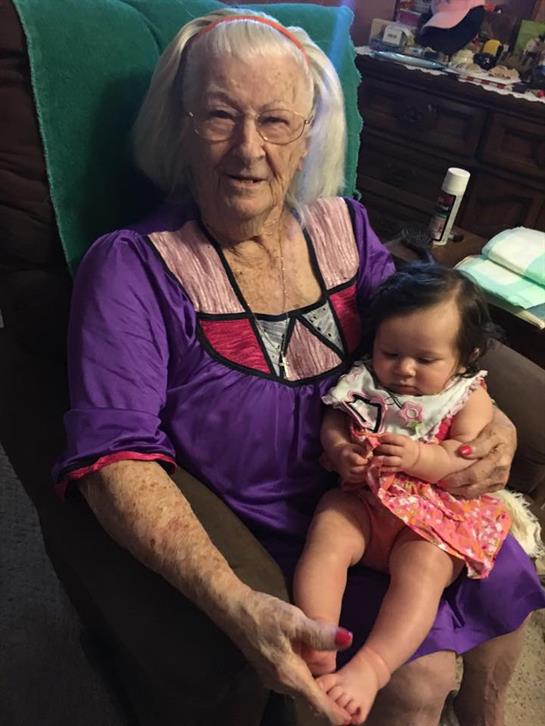 An elderly woman smiles in a chair while cuddling a baby girl on her lap.