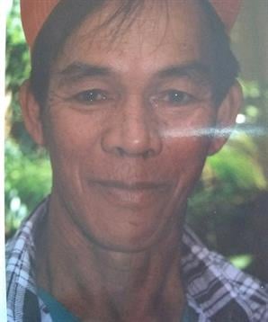 A man is smiling warmly while wearing a plaid shirt, surrounded by lush greenery on a clear day.