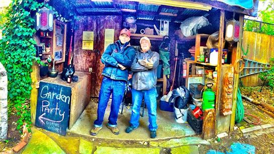 Two people proudly stand before a small workshop, showcasing their friendship and space.