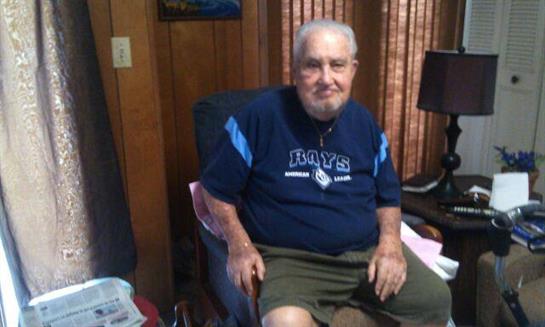 An elderly man sits comfortably in a home office, wearing summer clothes and smiling contently.
