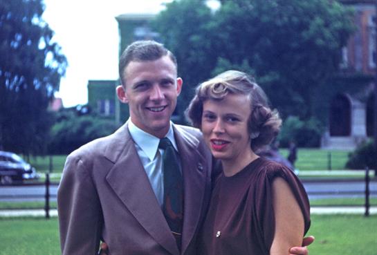 A couple smiles warmly at the camera while standing in a park setting surrounded by greenery.