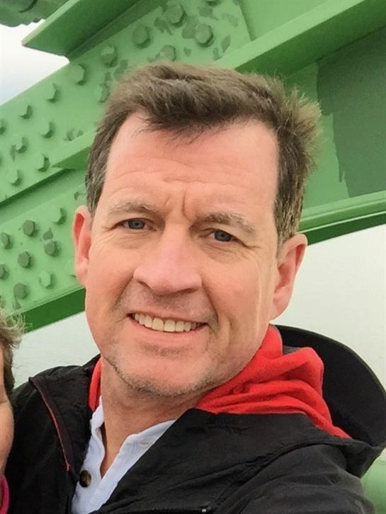 Man wearing a red jacket smiles against an iconic green structure under clear blue skies.