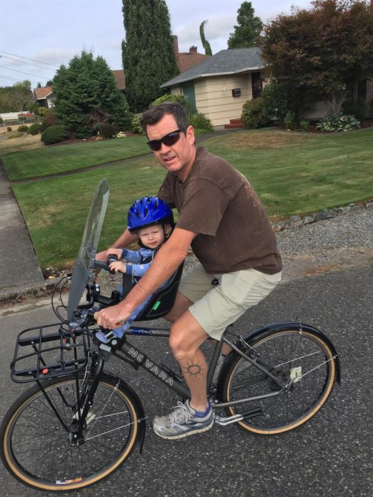 A father rides a bike with his young son, who is wearing a helmet, on a residential street.