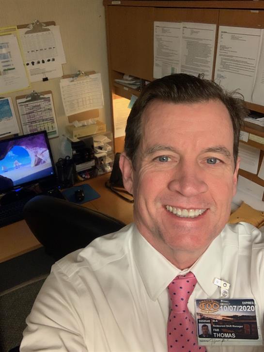 An office worker takes a cheerful selfie at his tech-filled desk.