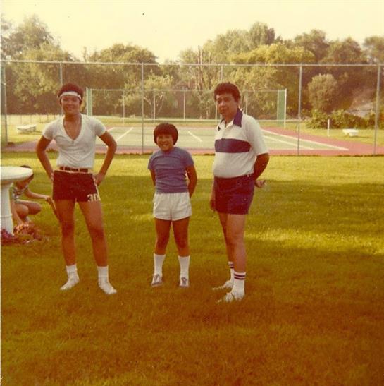 Three individuals stand on green grass beside tennis courts on a sunny day, smiling together.
