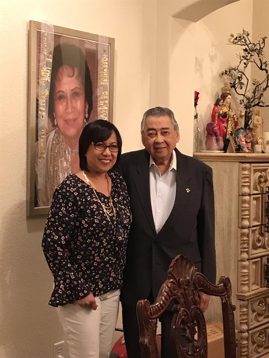 Two individuals smile while standing close to each other in a warm living space.