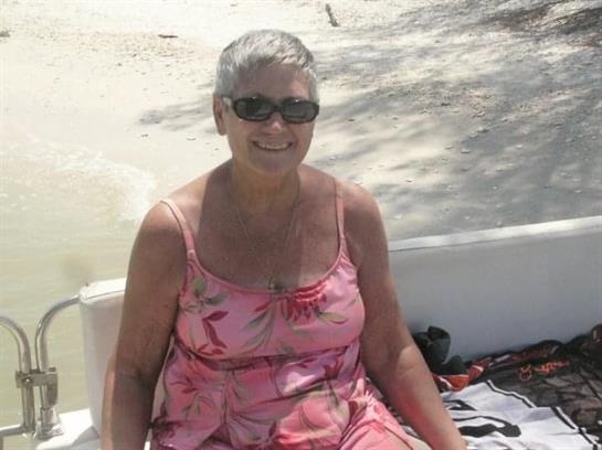 Woman with short grey hair and sunglasses smiles while sitting in a boat on the beach.