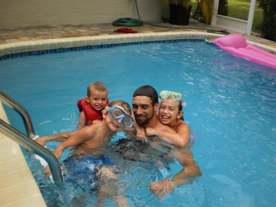 A man and three children play and swim in a backyard pool on a sunny summer day.