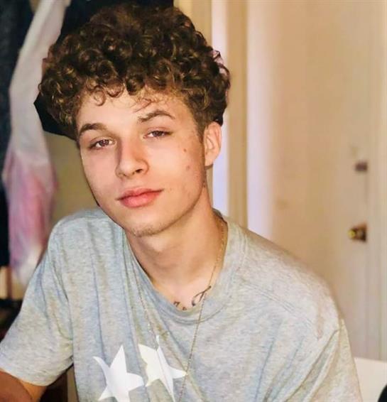 A young individual with curly hair poses in a cozy indoor setting, showcasing a laid-back vibe.