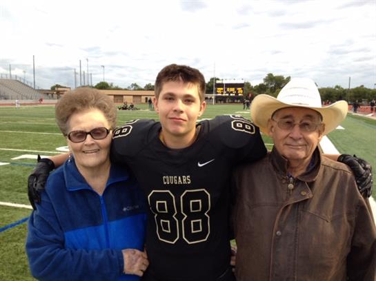 A young football player poses with his grandparents on the field after a game, smiling proudly.