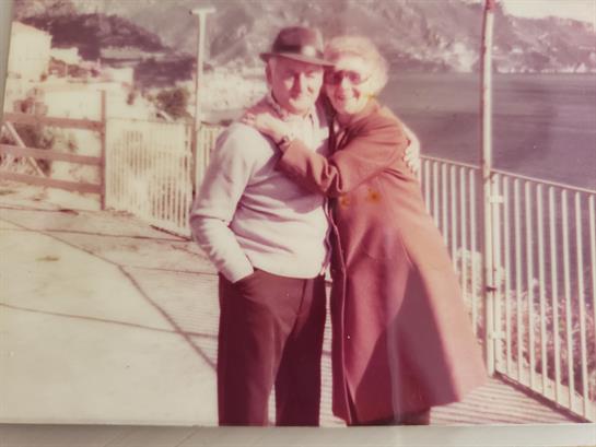 A couple stands close together on a coastal walkway, enjoying a romantic moment at sunset.