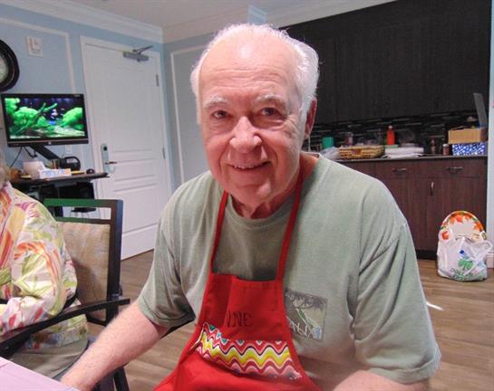 An elderly man cooks in a warm kitchen, sharing joy and laughter with family members around him.