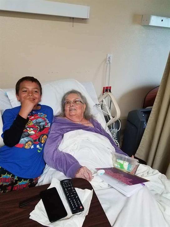 A young boy sits beside his grandmother in a hospital room, both smiling warmly.