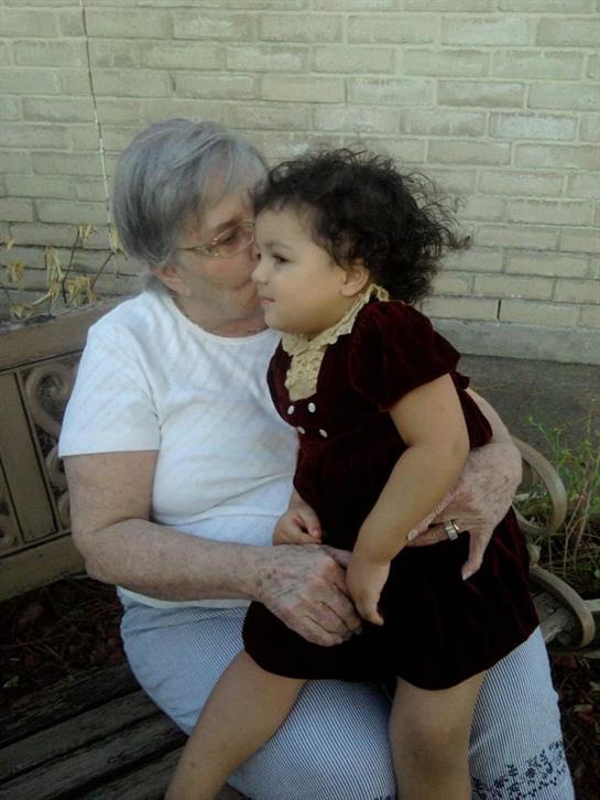 An elderly woman tenderly kisses a young girl sitting on her lap in a garden.