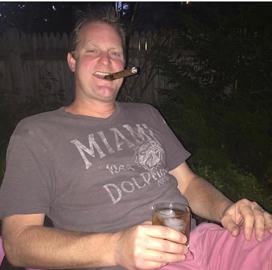 A man sits comfortably in the backyard at night, savoring a cigar and drink.