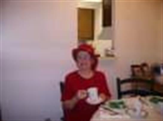 A senior woman sits at a dining table holding a tea cup, surrounded by cheerful decor and snacks.