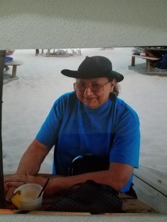 An older individual wearing a blue shirt and hat relaxes at a beachside table, sipping coffee.