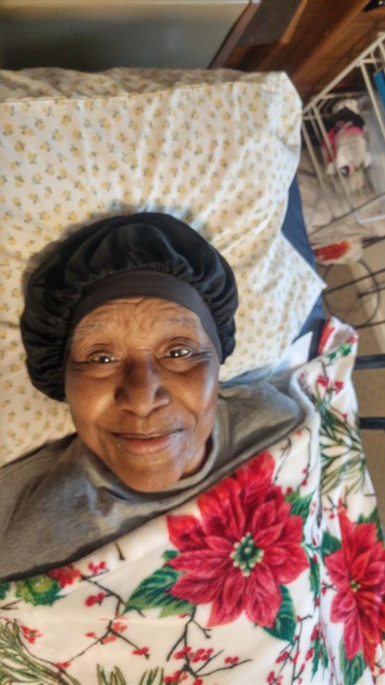 A smiling elderly woman lies on a bed, wrapped in a floral blanket, enjoying a moment of relaxation.