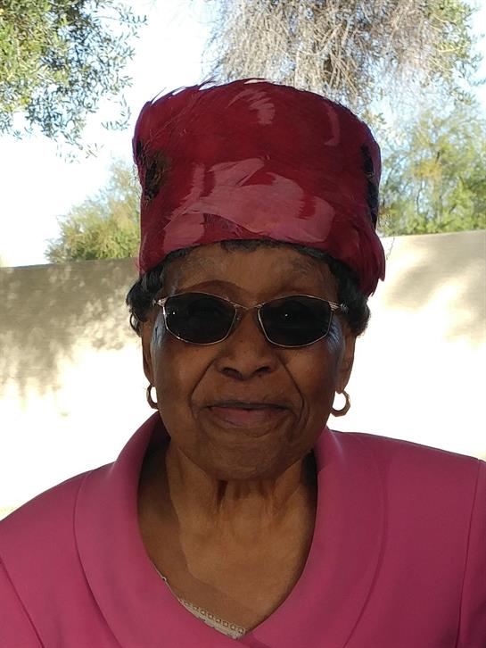 Joyful elderly woman stands outdoors wearing sunglasses and a vibrant pink headwrap, exuding warmth.
