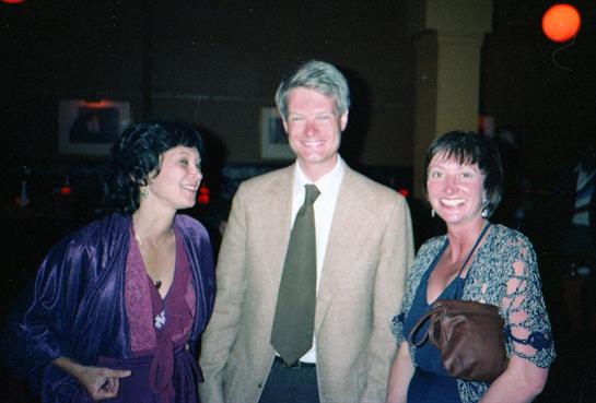 Three individuals stand close together, smiling and engaging in conversation at an evening event.