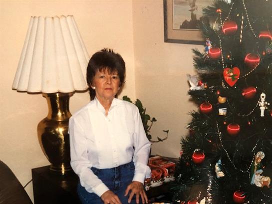 An elderly woman stands by a beautifully decorated Christmas tree in a cozy living room.