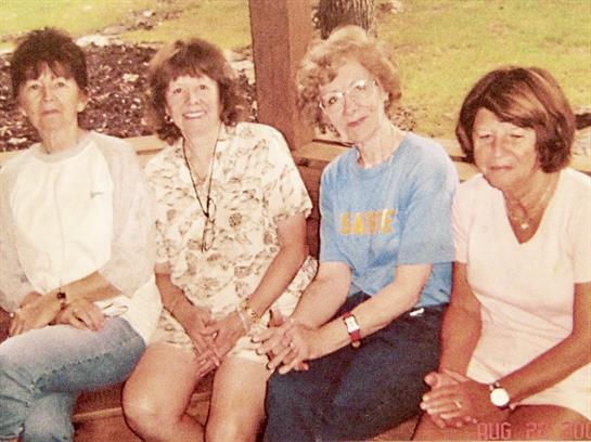 Four women are seated together on a wooden bench, smiling and chatting in a sunny outdoor setting.